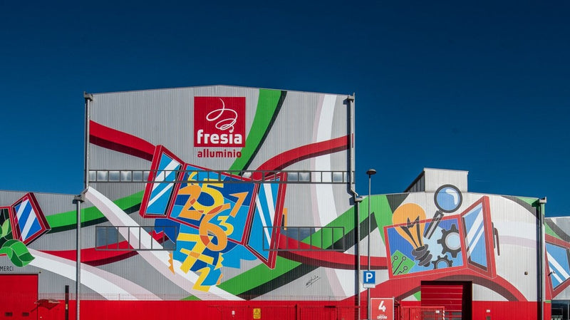 Murale colorato con motivi geometrici e iconografia artistica sulla facciata di un edificio industriale, rappresentando l'impegno di Mycrom.art per l'innovazione e la sostenibilità.