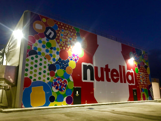 Una vibrante facciata murale notturna della Nutella con un design colorato di cerchi e bolle in varie dimensioni e colori brillanti che decorano estesamente la parete di un edificio industriale, illuminato vivacemente da faretti esterni.